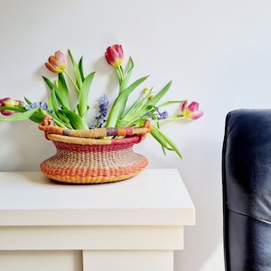 Peut inclure: Un panier tissé avec un motif rayé coloré contient un bouquet de tulipes roses et de fleurs violettes. Le panier est posé sur une cheminée blanche.