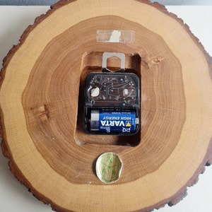 May include: The back of a wooden clock with a round, tree-trunk-shaped face. The clock is powered by a single AA battery, which is visible in the image. The battery is a VARTA brand, high energy battery. The clock has a small, round sticker on the bottom with the letters "F H K" on it.