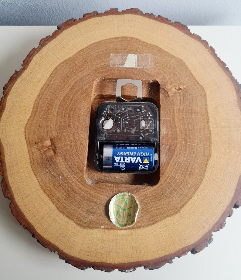 May include: The back of a wooden clock with a round, tree-trunk-shaped face. The clock is powered by a single AA battery, which is visible in the image. The battery is a VARTA brand, high energy battery. The clock has a small, round sticker on the bottom with the letters "F H K" on it.