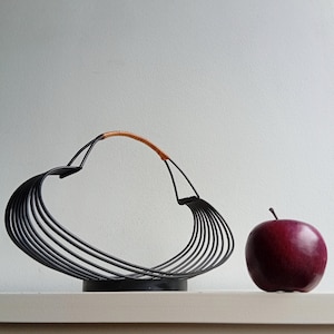 May include: Black metal fruit basket with a brown handle and a red apple. The basket has a heart-shaped design with multiple curved bars. The apple is a deep red color and sits next to the basket.