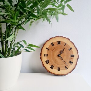 May include: A wooden clock with Roman numeral markings on a natural wood slice. The clock has black hands and is set against a white background.