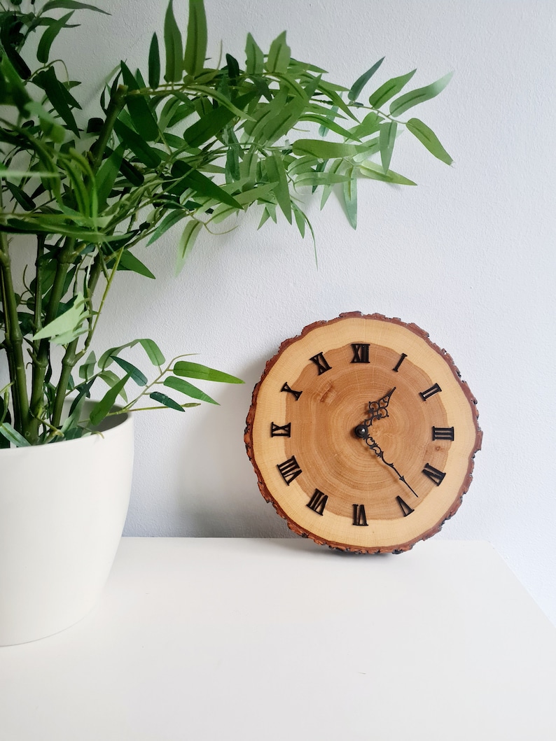 May include: A wooden clock with Roman numeral markings on a natural wood slice. The clock has black hands and is set against a white background.