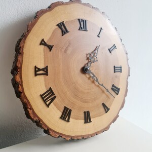 May include: A wooden clock with Roman numeral markings on a natural wood slice. The clock face is made of wood with a natural grain pattern. The clock hands are black metal.