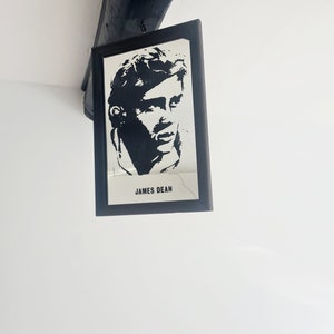 May include: Black and white framed print of James Dean's face. The print is in a black frame and hangs on a white wall.