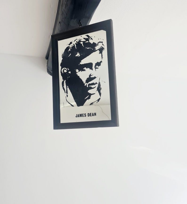May include: Black and white framed print of James Dean's face. The print is in a black frame and hangs on a white wall.