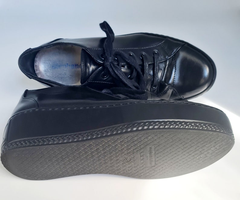 Puede incluir: Zapatillas de plataforma de cuero negro con suela gruesa de goma y cierre de cordones. La suela tiene un patr&oacute;n texturizado y las palabras "made in Italy" est&aacute;n impresas en la parte inferior.