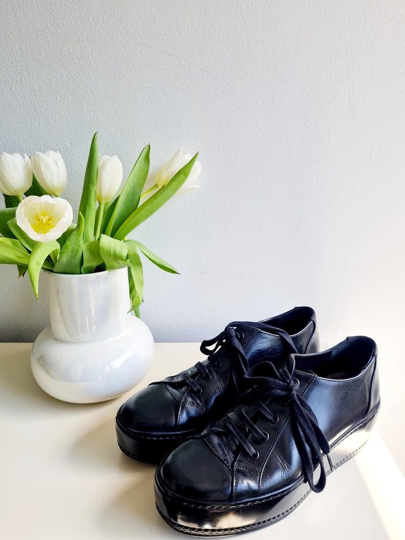 Puede incluir: Un par de zapatillas de plataforma de cuero negro con cordones blancos. Las zapatillas est&aacute;n sobre una superficie blanca junto a un jarr&oacute;n blanco con tulipanes blancos.