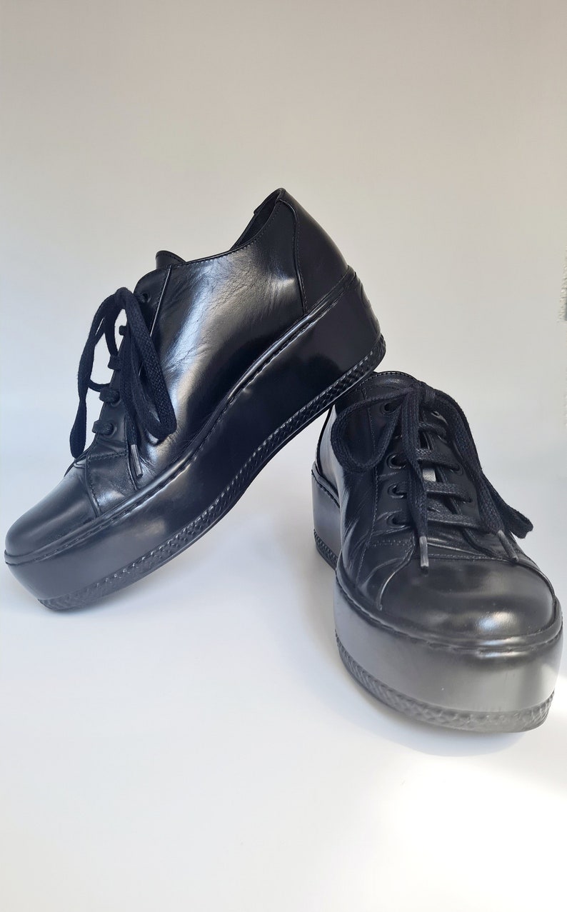 Puede incluir: Par de zapatillas de plataforma de cuero negro con cierre de cordones. Los zapatos tienen una suela gruesa y robusta.