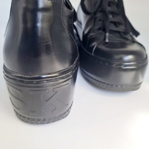 Puede incluir: Un par de zapatillas de plataforma de cuero negro con cierre de cordones. Las zapatillas tienen una suela de plataforma gruesa y el nombre de la marca "SK" est&aacute; estampado en la parte posterior del zapato izquierdo.