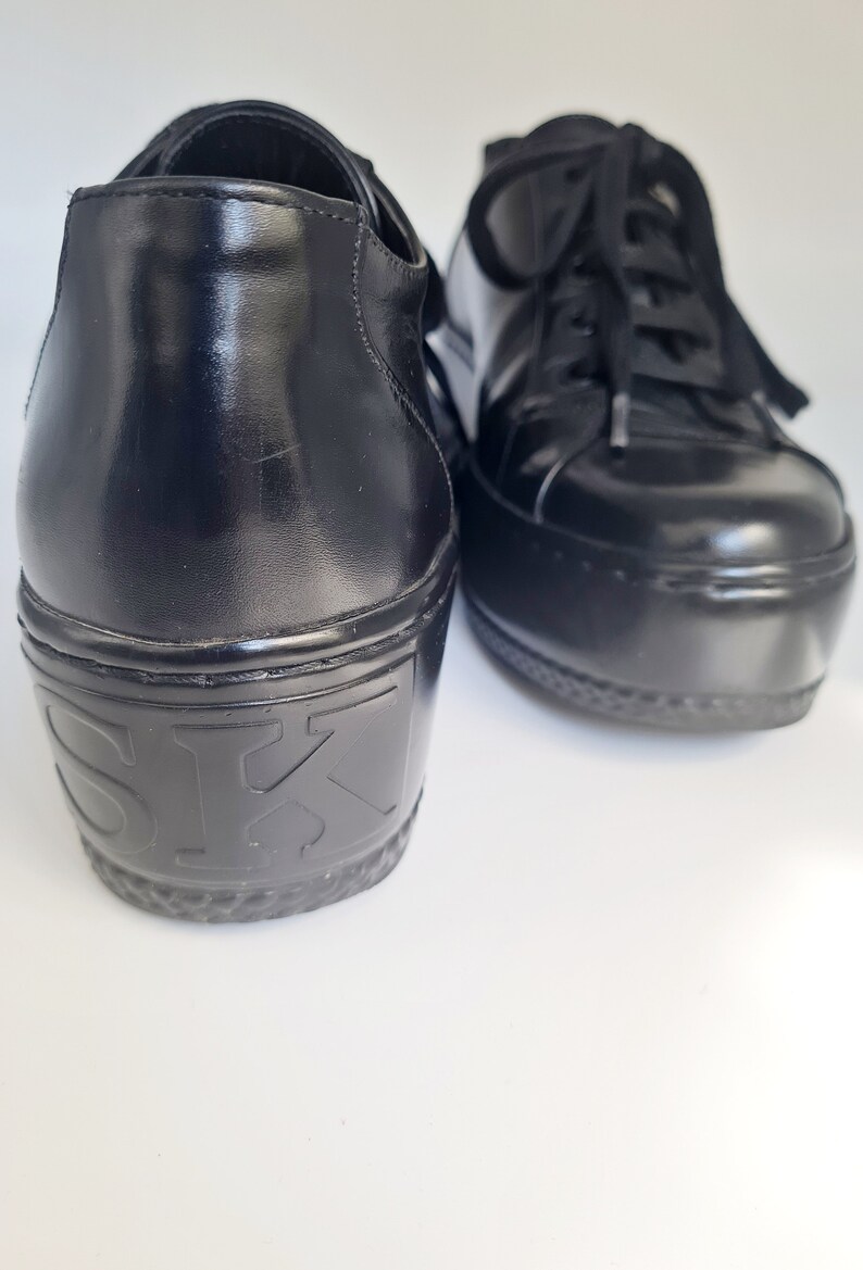 Puede incluir: Un par de zapatillas de plataforma de cuero negro con cierre de cordones. Las zapatillas tienen una suela de plataforma gruesa y el nombre de la marca "SK" est&aacute; estampado en la parte posterior del zapato izquierdo.
