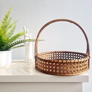 May include: A round, woven wicker basket with a single handle. The basket is brown and has a natural finish. It is sitting on a white surface.
