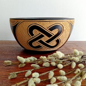 May include: A hand-carved wooden bowl with a black interior and a light brown exterior. The bowl features a Celtic knot design carved into the side. The bowl is sitting on a wooden surface with a sprig of pussy willow.