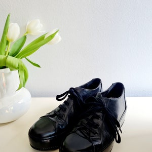 Puede incluir: Un par de zapatillas de plataforma de cuero negro con cordones negros. Las zapatillas tienen un acabado brillante y una puntera redondeada.