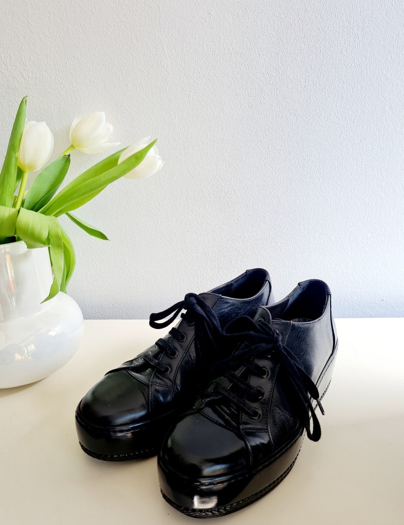 Puede incluir: Un par de zapatillas de plataforma de cuero negro con cordones negros. Las zapatillas tienen un acabado brillante y una puntera redondeada.