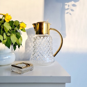 Puede incluir: Una jarra de vidrio transparente con tapa y asa doradas. La jarra tiene un patrón de diamante y está sentada sobre una mesa blanca con un pequeño cenicero de vidrio transparente.