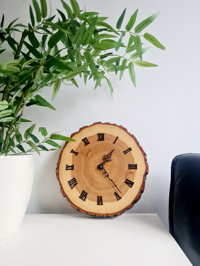 May include: A wooden clock with Roman numeral markings on a natural wood slice. The clock hands are black and the clock is hanging on a white wall.