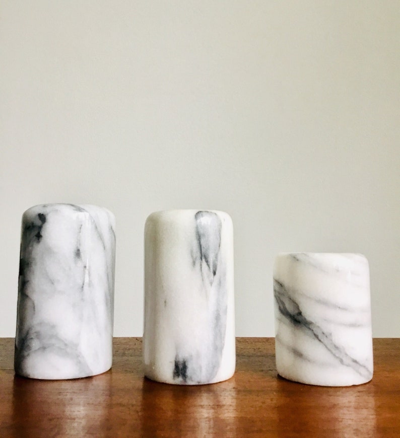 May include: Three white marble cylindrical containers with gray veining. The containers are arranged in a row from largest to smallest on a wooden surface.