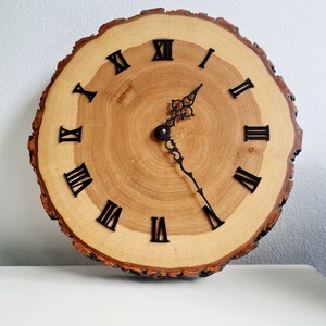 May include: A wooden clock with Roman numeral markings on a natural wood slice. The clock has black hands and a decorative design on the minute hand.