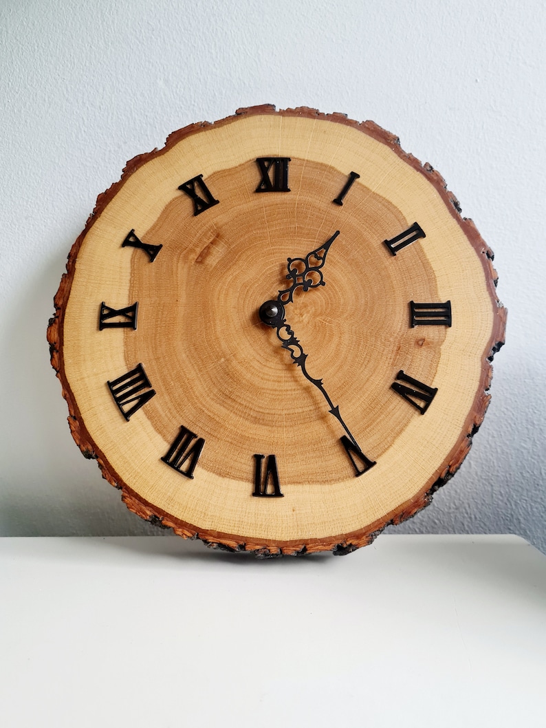May include: A wooden clock with Roman numeral markings on a natural wood slice. The clock has black hands and a decorative design on the minute hand.