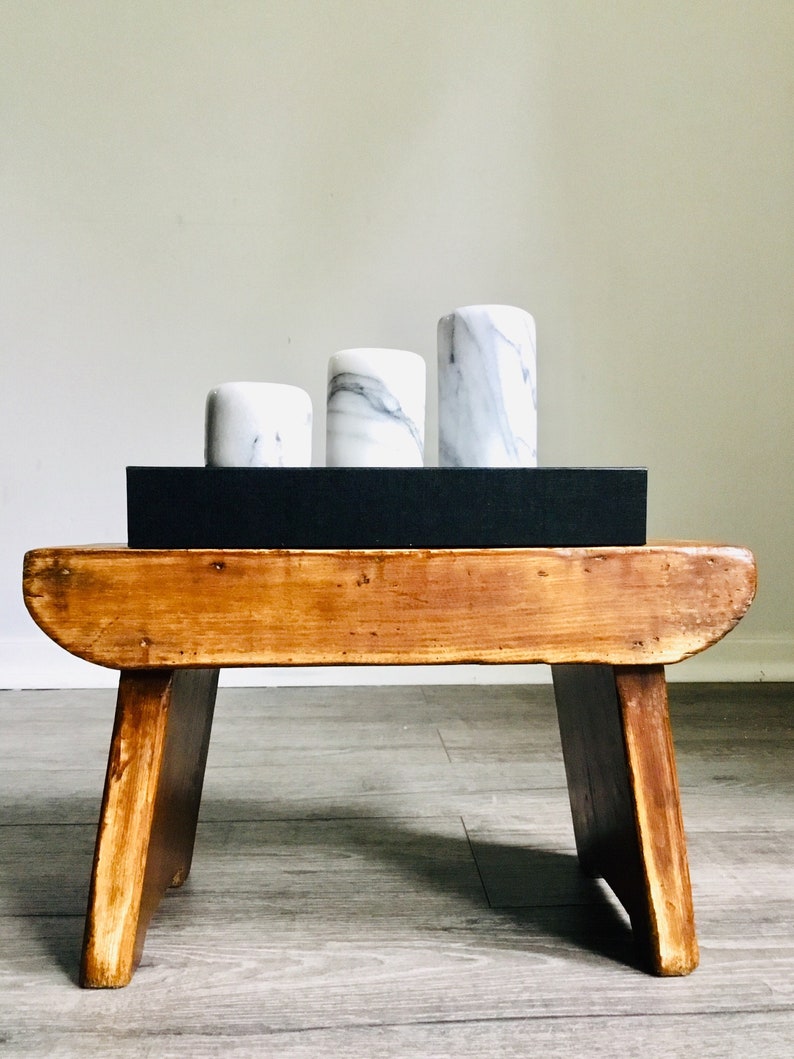 May include: Three white marble cylinders of varying sizes are stacked on a black rectangular tray, which is placed on a wooden stool with a natural finish.