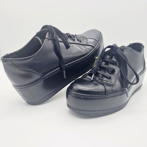 Puede incluir: Un par de zapatillas de plataforma de cuero negro con cordones. Las zapatillas tienen una suela de plataforma gruesa y una puntera redonda.