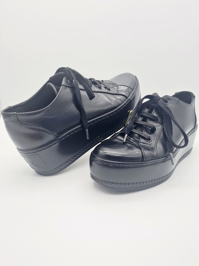 Puede incluir: Un par de zapatillas de plataforma de cuero negro con cordones. Las zapatillas tienen una suela de plataforma gruesa y una puntera redonda.