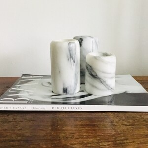 May include: Three white marble candle holders with gray veining. The candle holders are arranged in a row on a magazine.