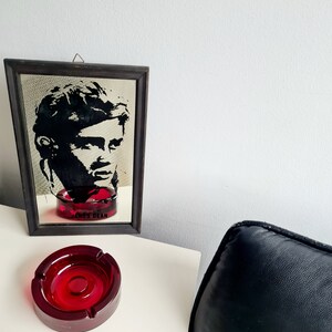 May include: A red glass ashtray with a black framed mirror behind it. The mirror has a black and white image of James Dean with the text "James Dean" below the image.
