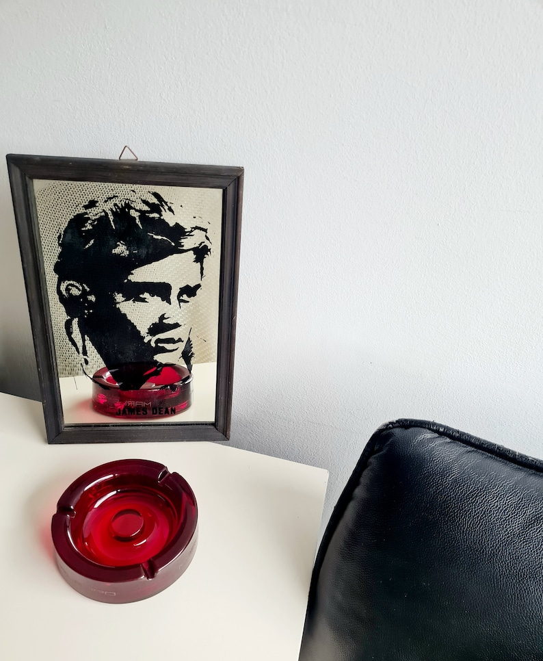 May include: A red glass ashtray with a black framed mirror behind it. The mirror has a black and white image of James Dean with the text "James Dean" below the image.