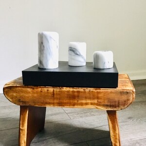 May include: Three white marble candle holders of varying sizes are arranged on a black rectangular tray. The tray is placed on a wooden stool with a rustic finish.