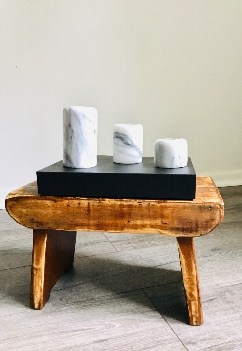 May include: Three white marble candle holders of varying sizes are arranged on a black rectangular tray. The tray is placed on a wooden stool with a rustic finish.