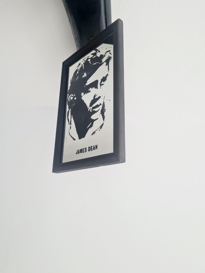 May include: Black and white framed print of James Dean, a famous actor. The print is in a black frame and is hanging on a white wall.