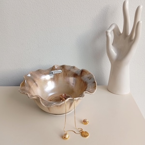 Pode incluir: Uma tigela decorativa de cerâmica bege e castanha com borda ondulada, contendo joias. Um colar e brincos dourados são exibidos próximos. À direita, há uma escultura de mão de cerâmica branca.