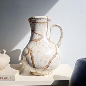 May include: A white ceramic vase with a brown crackle glaze and a handle. The vase is sitting on a white table.