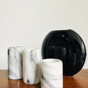 May include: Three white marble candle holders with black veining, next to a black ceramic vase with a round, slightly flattened shape.