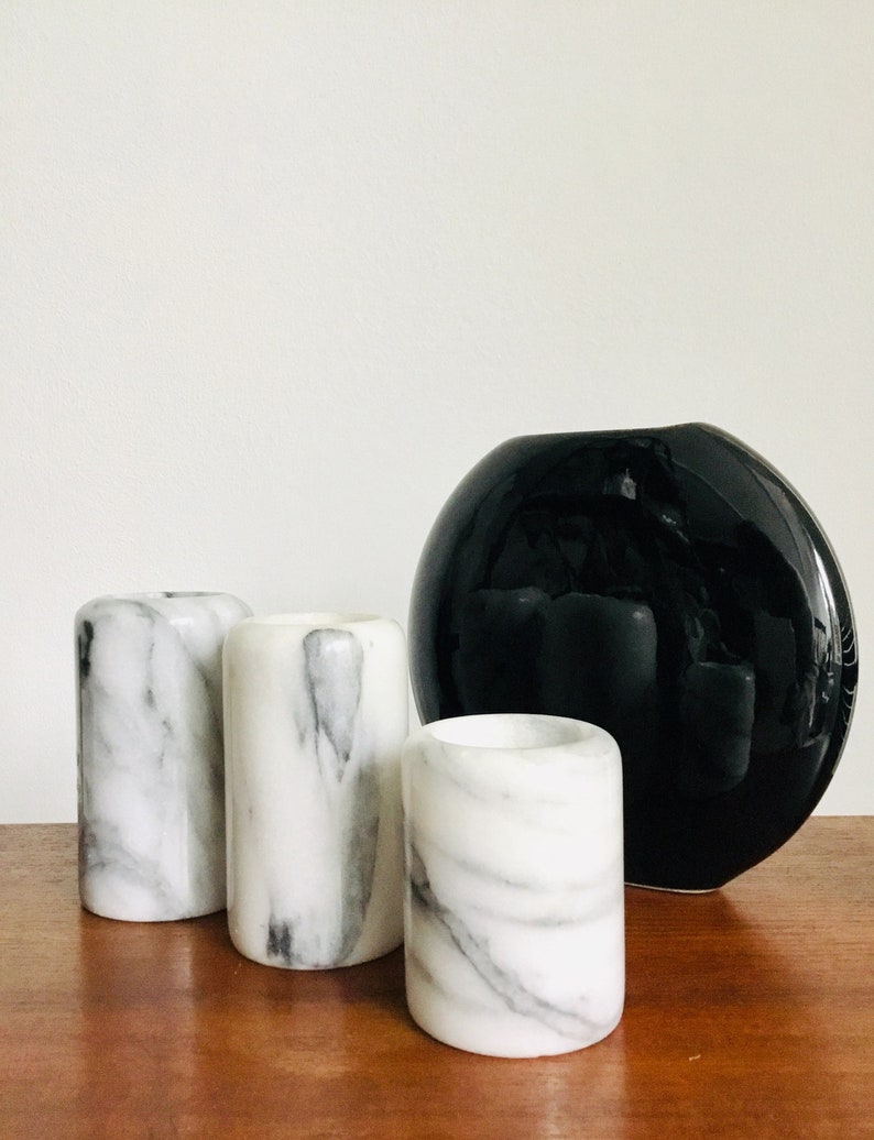 May include: Three white marble candle holders with black veining, next to a black ceramic vase with a round, slightly flattened shape.