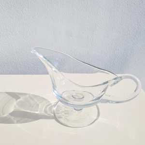 Puede incluir: Una salsera de vidrio transparente con asa y base de pedestal. La salsera está vacía y tiene una ligera curva en el pico.