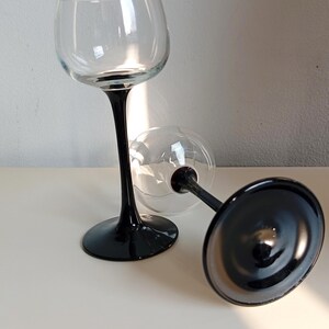 May include: Two clear wine glasses with black stems and bases. One glass stands upright, while the other lies on its side. The glasses have a classic, elegant design, suitable for serving wine.