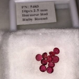 May include: Ten Burmese Red Ruby Round gemstones, each 2.5 mm in diameter, are displayed on a white surface. The gemstones are a deep red color and are round in shape. A label in the background reads "PN: 5483 10pcs 2.5 mm Burmese Red Ruby Round."