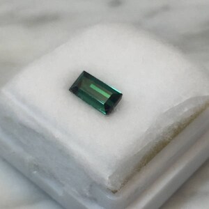 May include: A rectangular, emerald-cut gemstone with a deep teal color and a reflective surface. The gem is set on a white, textured surface, suggesting a presentation for jewelry or gemstones. The cut of the stone allows light to reflect, creating a vibrant appearance.