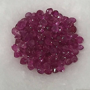 May include: A pile of small, round, pink ruby gemstones. The rubies are faceted and have a shiny, polished finish.