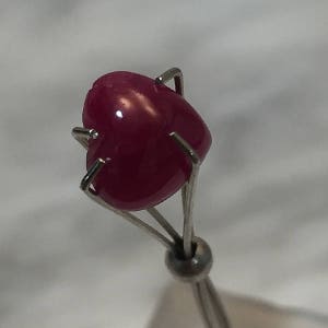 May include: A heart-shaped ruby gemstone held in a metal clamp. The ruby is a deep red color and has a smooth, polished surface.