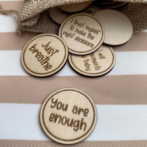 May include: A selection of wooden affirmation tokens with motivational phrases. The round tokens have a light brown border and feature phrases such as "Just breathe" and "You are enough" in a simple font. They are spilling out of a small hessian bag.
