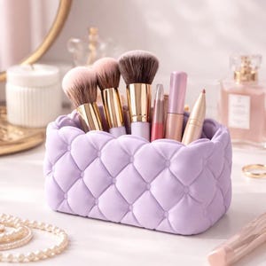 May include: A lavender quilted makeup brush holder filled with various makeup brushes and cosmetic products. The holder has a rectangular shape with rounded corners and a diamond pattern. Gold-toned brush handles and pink and brown brushes are visible.