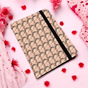 Puede incluir: Un porta pasaporte con un patrón repetido de la cara de una persona. El porta pasaporte está hecho de un material suave y tiene una banda elástica negra.