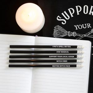 May include: A collection of black pencils with white erasers, each inscribed with a different phrase: "I PUT A SPELL ON YOU", "STAY MAGICAL", "SUPPORT YOUR LOCAL COVEN", "WITCH VIBES", and "RESTING WITCH FACE". A lit white candle sits above the pencils on an open notebook.