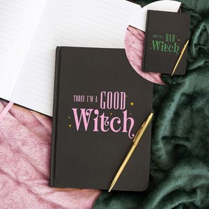 May include: Black notebook with the text "Today I'm a Good Witch" in pink script and a gold pen. A smaller notebook with the text "Today I'm a Bad Witch" is also visible. The notebooks are on a pink and green surface.