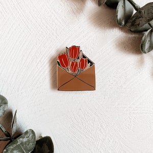 Puede incluir: Un pin de esmalte dorado con un sobre marr&oacute;n con un ramo de tulipanes rojos en su interior.