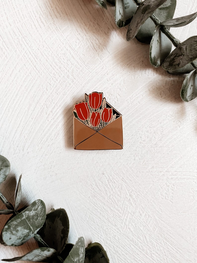 Puede incluir: Un pin de esmalte dorado con un sobre marr&oacute;n con un ramo de tulipanes rojos en su interior.