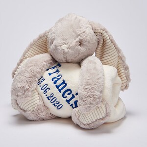 May include: A plush gray bunny holding a white blanket with blue embroidery. The blanket is personalized with the name "Francis" and the date "13.06.2020".